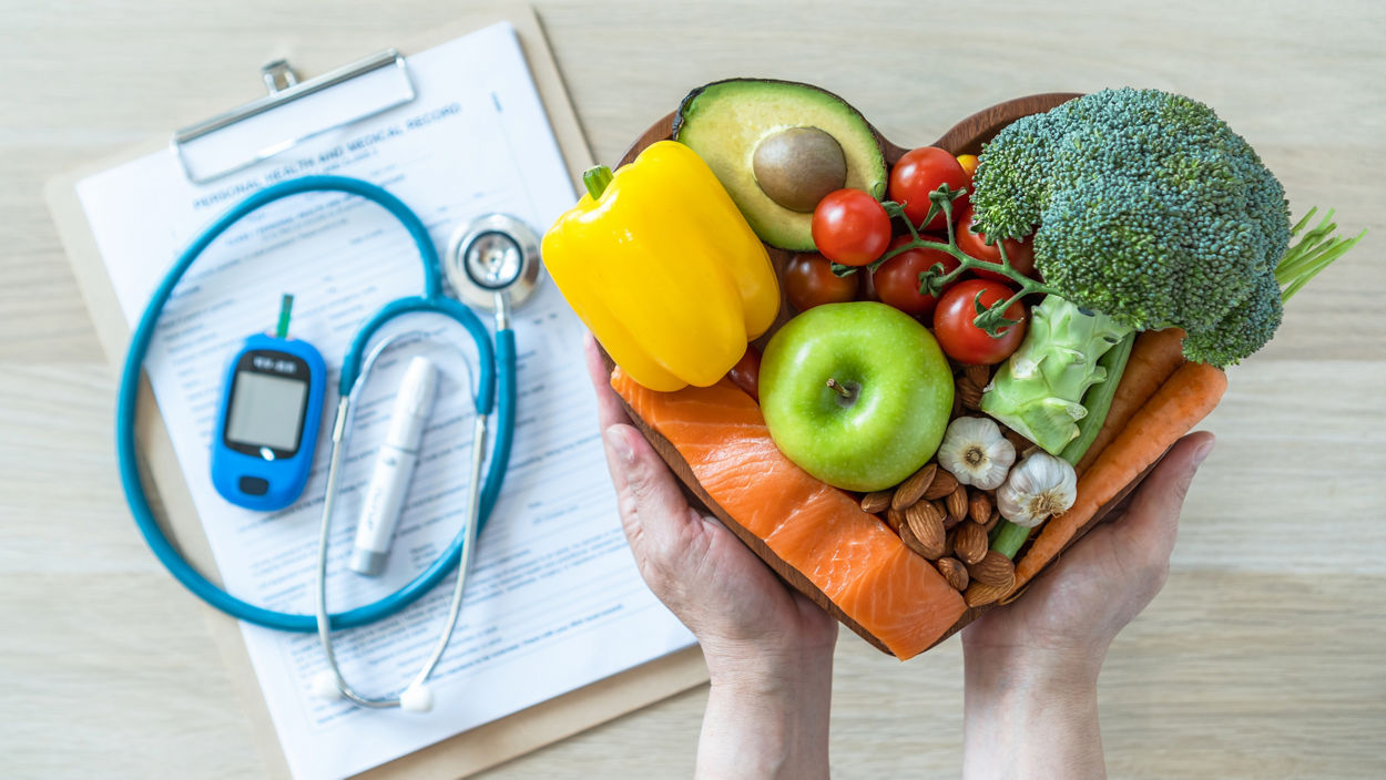 Person holding a heart-shaped bowl with fruits and vegetables with a stethoscope, glucose meter, and clipboard in the background