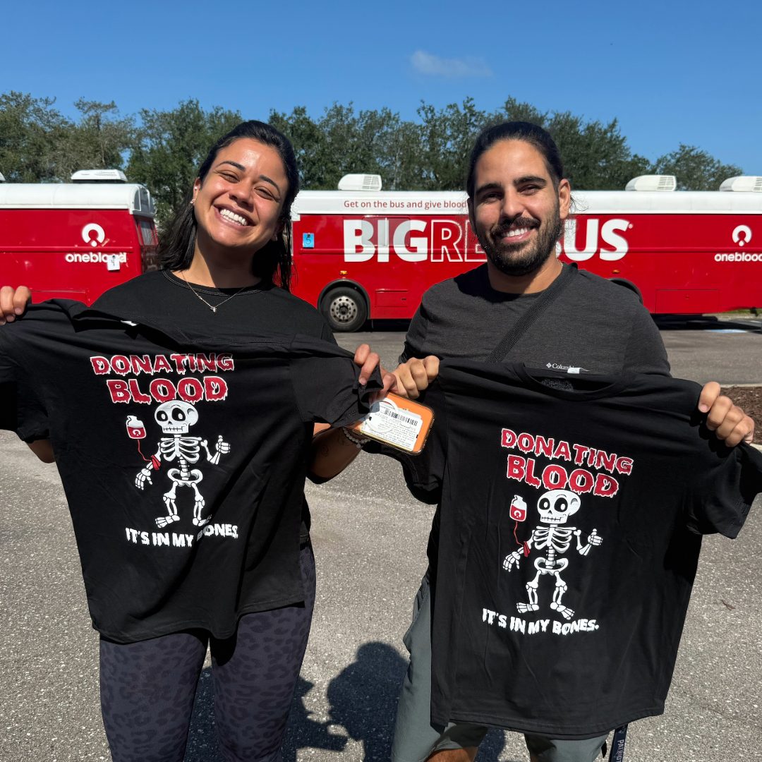 Two people hold up black Halloween T-shirts featuring a skeleton holding a blood bag with the words “Donating Blood, It’s in My Bones.” A red OneBlood Big Red Bus is parked behind them.